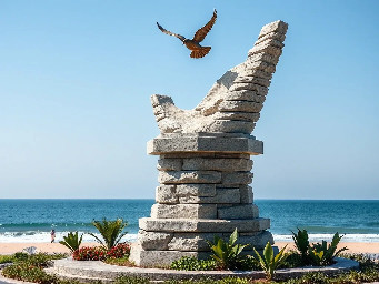 A 5-meter tall majestic sculpture made of stacked natural stones, featuring a prominent hexagonal base structure with a 6-meter diameter. At the very top, a beautifully sculpted, large-sized bronze dove is captured in mid-flight, symbolizing peace and freedom. The monument is situated in a public park right along the scenic beachfront of Sihanoukville, Cambodia. The base is surrounded by a lush coastal garden with native salt-tolerant plants and beach vegetation. Bright daylight, clear blue sky, soft ocean waves in the background, photorealistic, architectural rendering style, highly detailed stone and polished metal textures, 8k resolution.