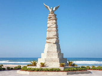 A 5-meter tall majestic sculpture made of stacked natural stones, featuring a hexagonal base structure with a 6-meter diameter. At the very top, a beautifully sculpted stone dove is captured in mid-flight, symbolizing peace and freedom. The monument is situated in a public park right along the scenic beachfront of Sihanoukville, Cambodia. The base is surrounded by a lush coastal garden with native salt-tolerant plants and beach vegetation. Bright daylight, clear blue sky, soft ocean waves in the background, photorealistic, architectural rendering style, highly detailed stone textures, 8k resolution.