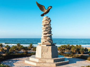 A 5-meter tall majestic sculpture made of stacked natural stones, featuring a prominent hexagonal base structure with a 6-meter diameter. At the very top, a beautifully sculpted, large-sized bronze dove is captured in mid-flight, symbolizing peace and freedom. The monument is situated in a public park right along the scenic beachfront of Sihanoukville, Cambodia. The base is surrounded by a lush coastal garden with native salt-tolerant plants and beach vegetation. Bright daylight, clear blue sky, soft ocean waves in the background, photorealistic, architectural rendering style, highly detailed stone and polished metal textures, 8k resolution.