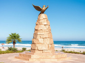 A 5-meter tall majestic sculpture made of stacked natural stones, featuring a prominent hexagonal base structure with a 6-meter diameter. At the very top, a beautifully sculpted, large-sized bronze dove is captured in mid-flight, symbolizing peace and freedom. The monument is situated in a public park right along the scenic beachfront of Sihanoukville, Cambodia. The base is surrounded by a lush coastal garden with native salt-tolerant plants and beach vegetation. Bright daylight, clear blue sky, soft ocean waves in the background, photorealistic, architectural rendering style, highly detailed stone and polished metal textures, 8k resolution.